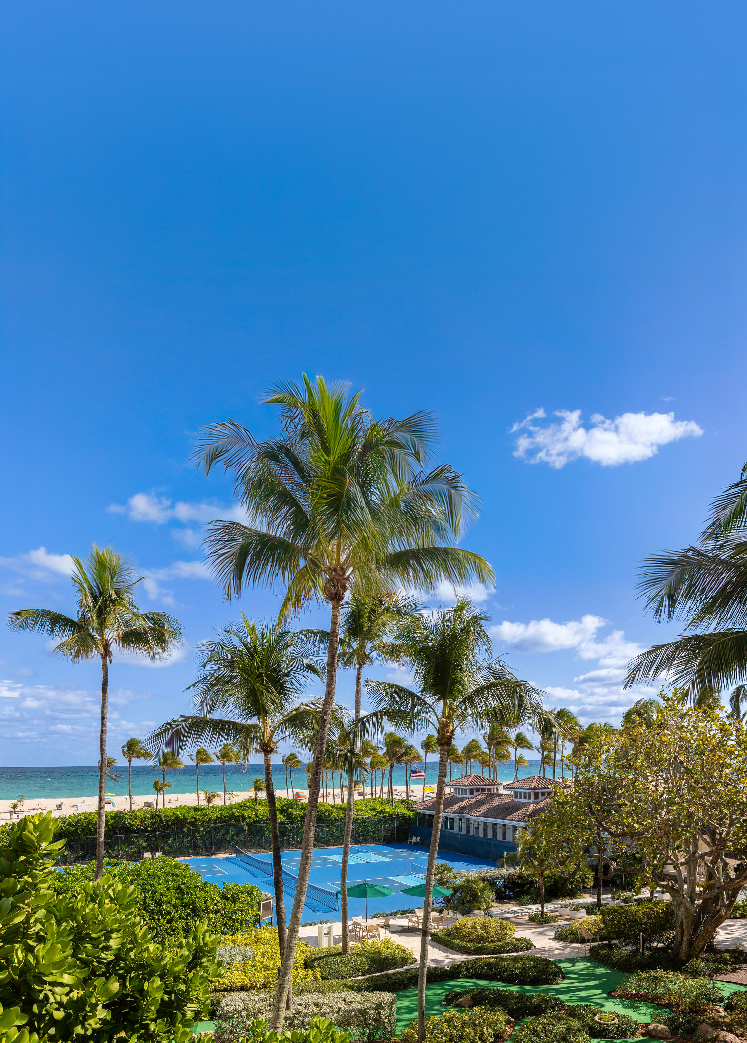 A beachfront view of a resort with palm trees, a tennis court, and a beach.
